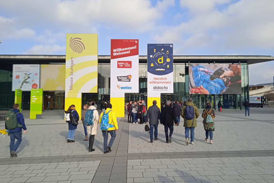 Auf dem Bild sieht man den großen Vorplatz der Messe Stuttgart mit verschiedenen Menschen, die in Richtung des Messeeingangs laufen. Man kann mehrere Schilder sehen, u. a. eines das noch einmal die didacta bewirbt.